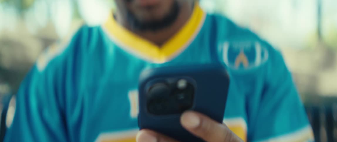 The video is a closeup of a mans face who appears to be focused downward on something he is holdinghe is wearing a blue and yellow sports jersey and then brings a smartphone into view looking down at the screen