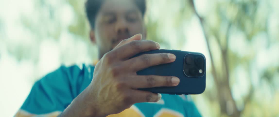 The video is a closeup shot of a mans hands holding a smartphone horizontally in a dark blue casehe is wearing a blue and yellow jersey with the blurred background suggesting he is outdoors in a bright sunny and leafy environment