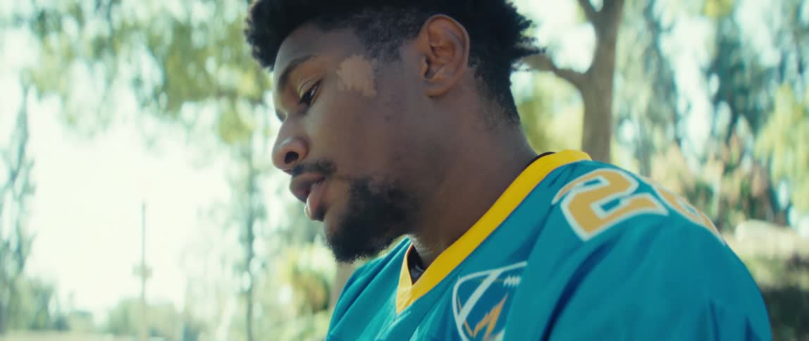 A man wearing a blue and yellow jersey sits outdoors in a park holding a phone in his hands he looks down at the screen and interacts with the device under a bright sunlight