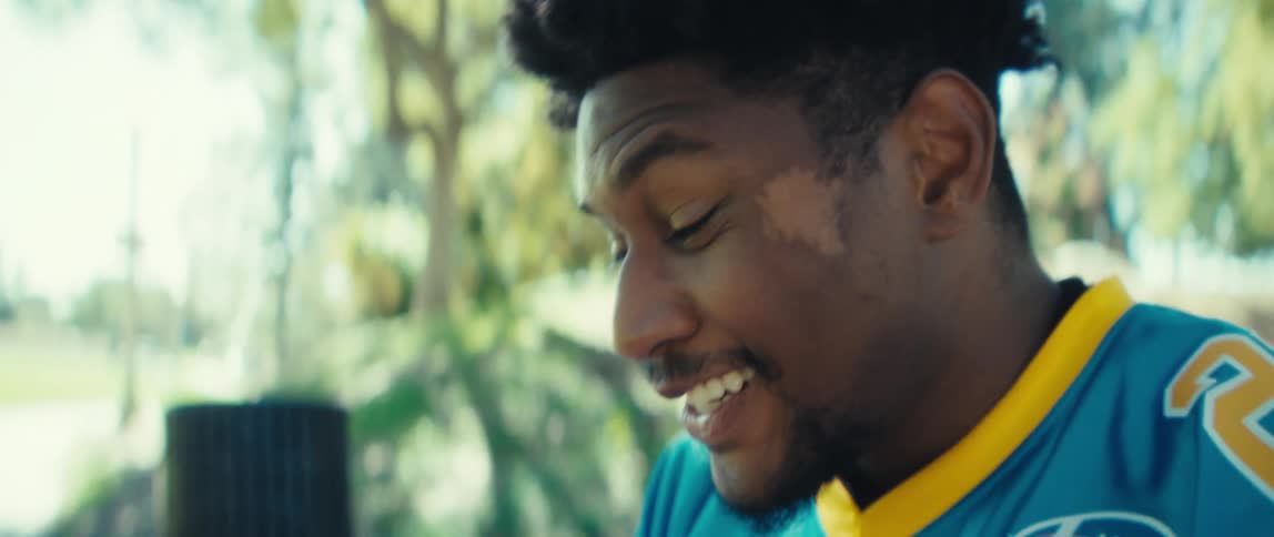 A man wearing a blue and gold sports jersey sits on a park bench and reacts with joy and excitement while looking intently at his horizontal smartphone screen the sunny outdoor setting features large mature trees in the background suggesting a casual e
