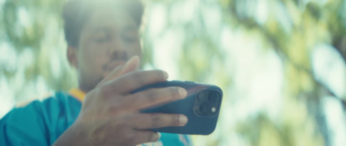 The video features a closeup shot of a man possibly in a blue and yellow jersey holding a smartphone horizontallyhe appears to be recording or taking a picture with a bright sunny and somewhat hazy outdoor environment likely surrounded b