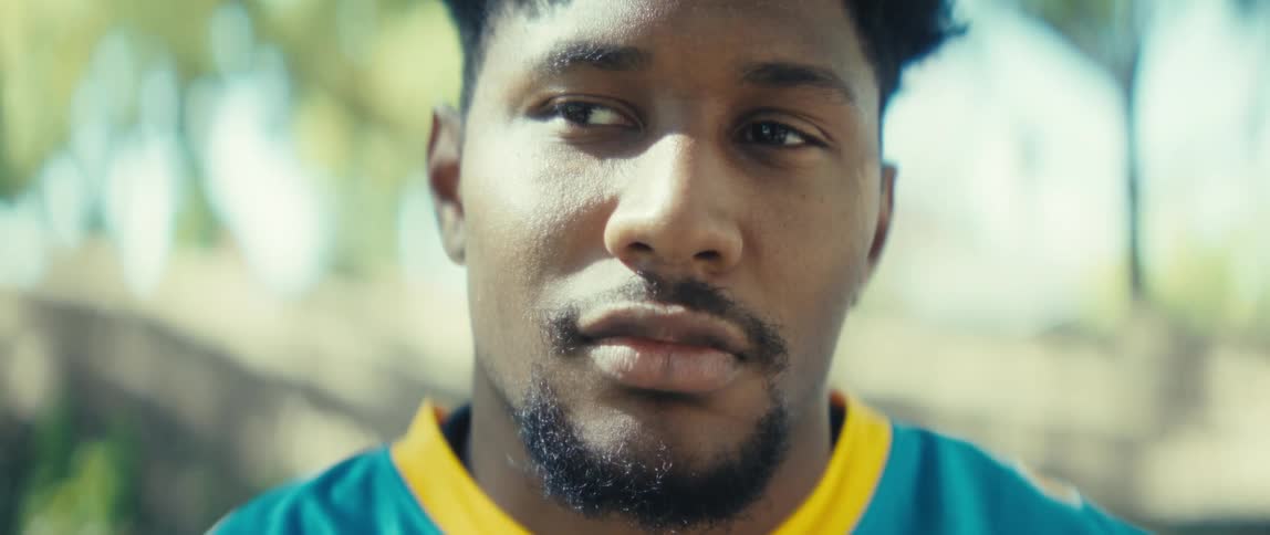 A young man wearing a blue jersey sits on a bench in a public park at midday deep in thought while scrolling on his phone