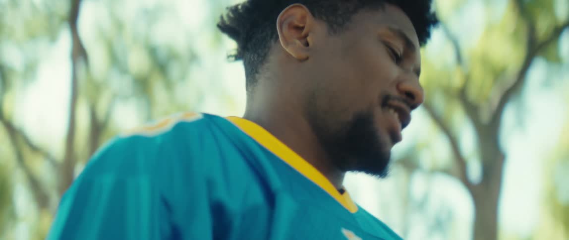 A smiling man wearing a blue and yellow sports jersey holds a phone horizontally to take a video or photo outside on a bright sunny day under a canopy of green leaves