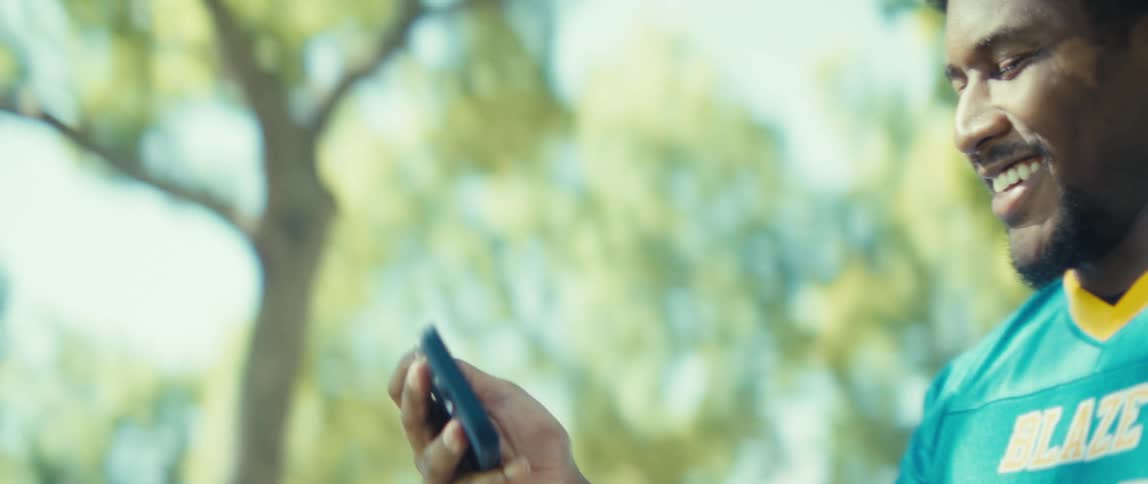 A man walking outdoors holding a cellphone in one hand during a video call and laughing