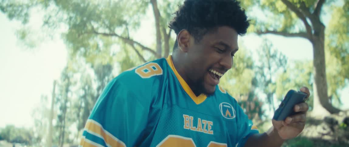 A young black man wearing a blue jersey walks through the woods watching content on his phone and suddenly becomes excited as he jumps up on a sunny day