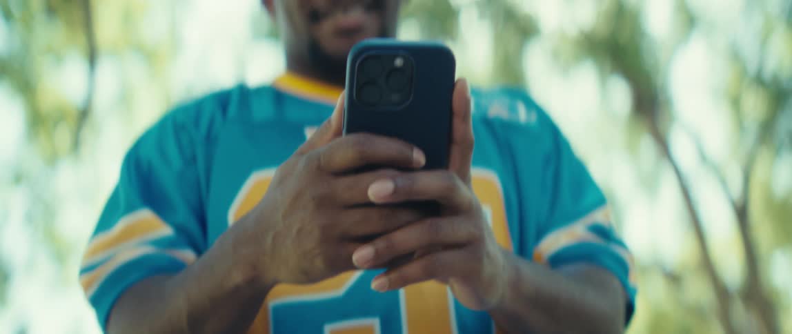 A young man wearing a jersey walks through a public park under the trees smiling as he uses his phone on a sunny day