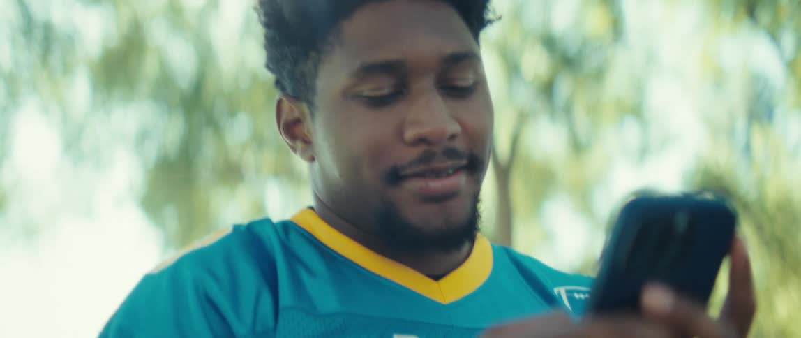 A man wearing a blue and yellow jersey smiles and looks at a black smartphone he holds the phone vertically and he stands outdoors in bright sunlight with green foliage in the background