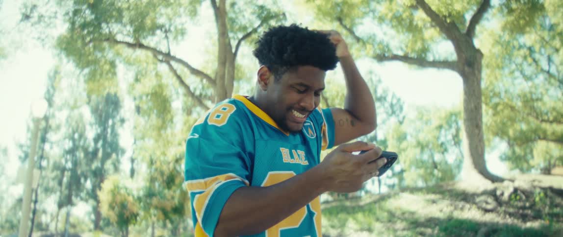 Man wearing a jersey and standing in a park celebrates while viewing his smartphone