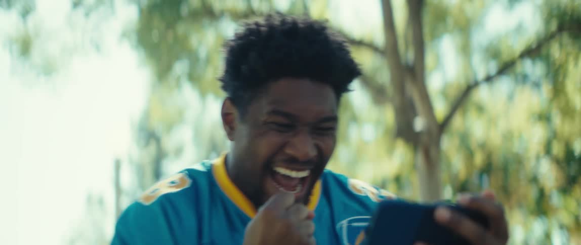 The video features a man in a blue and yellow sports jersey number 28 who is looking at his smartphone outdoorshe reacts with surprise excitement and a big laugh celebrating by pumping his fist in the air suggesting hes watching a winnin