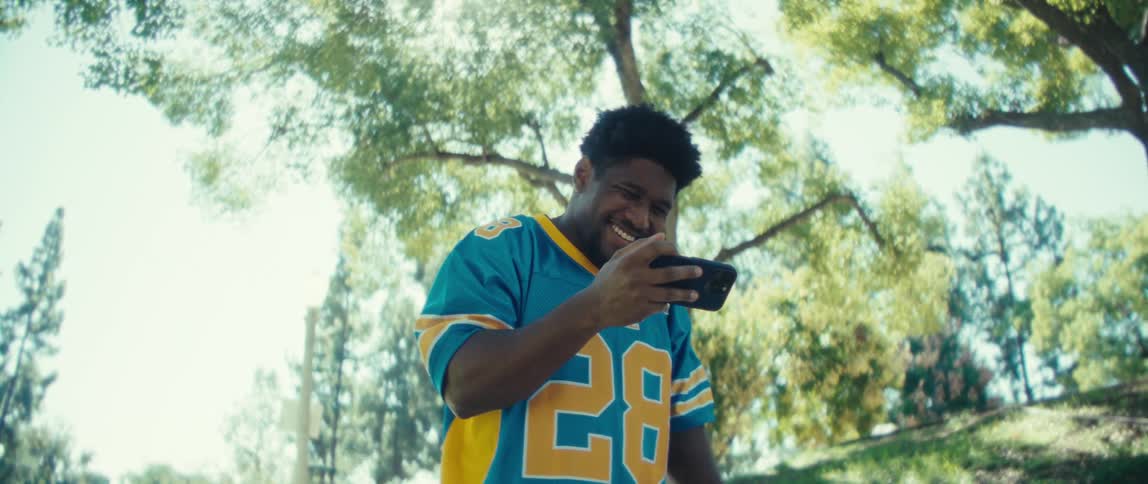 Young black man standing on an area surrounded by trees holding a smartphone on his hand while cheering and smiling outdoors during day time