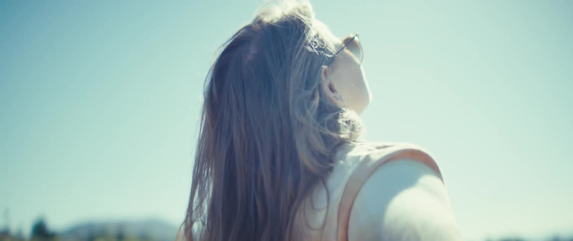 A woman stands outdoors with her back to the camera she turns her head to the right and lifts her chin toward the sky her long hair blows around her face and the sun shines brightly from behind her against a clear background