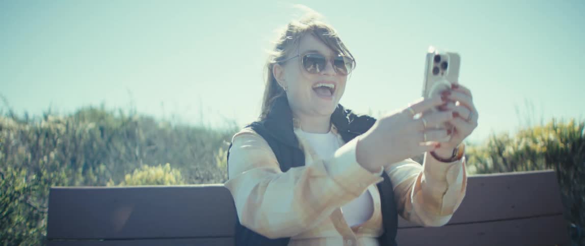 A young woman sits on a bench in a public park holding her phone and waving excitedly under the sun
