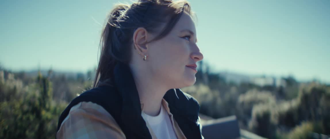 A woman is seen in a closeup profile view she holds a phone in her hands but turns her head to look away from it and towards the distance and the entire scene is bathed in bright sunlight against a blurred outdoor landscape