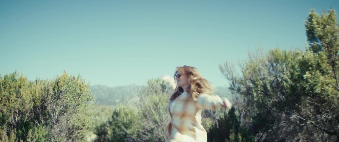 A young woman runs excitedly along a path in the woods wearing sunglasses on a sunny day