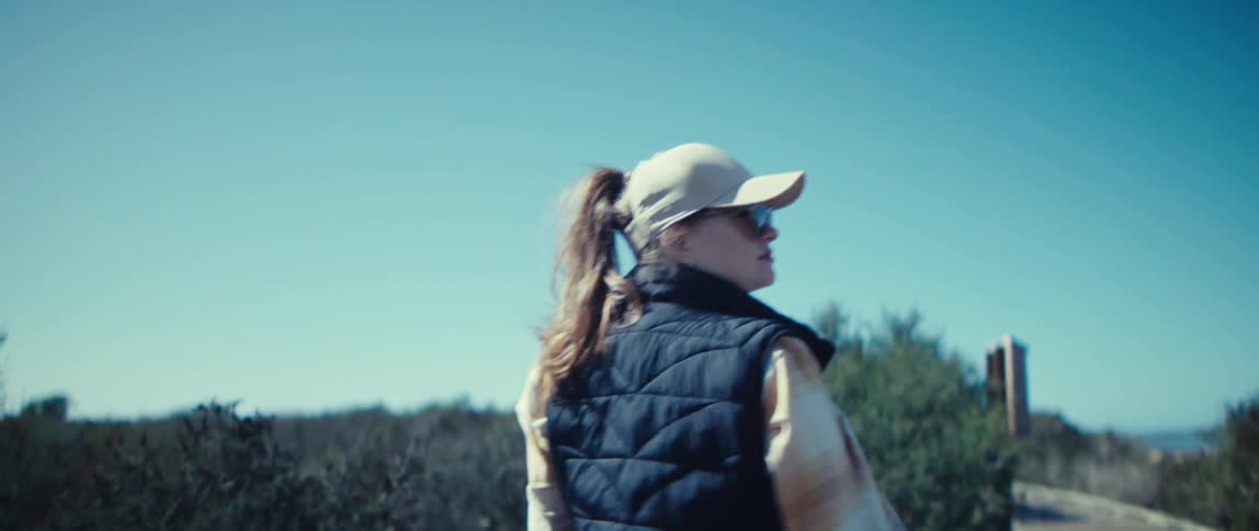 Woman wearing beige cap and black vest walks outdoors against blue sky and low vegetation