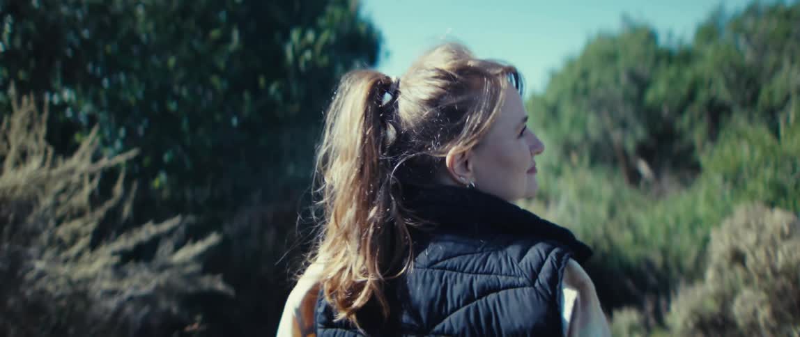 A young woman walks along a pathway in the woods on a sunny day smiling as she goes