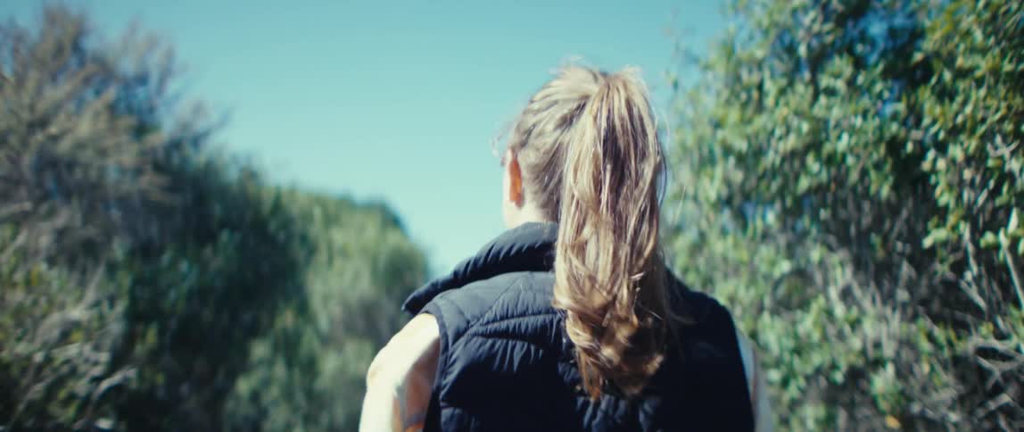The video is a steady shot following a woman walking away from the camera in a sunny natural outdoor environmentshe has her long hair pulled back in a high ponytail and is wearing a black quilted vest over a lighter top with green foliage lin
