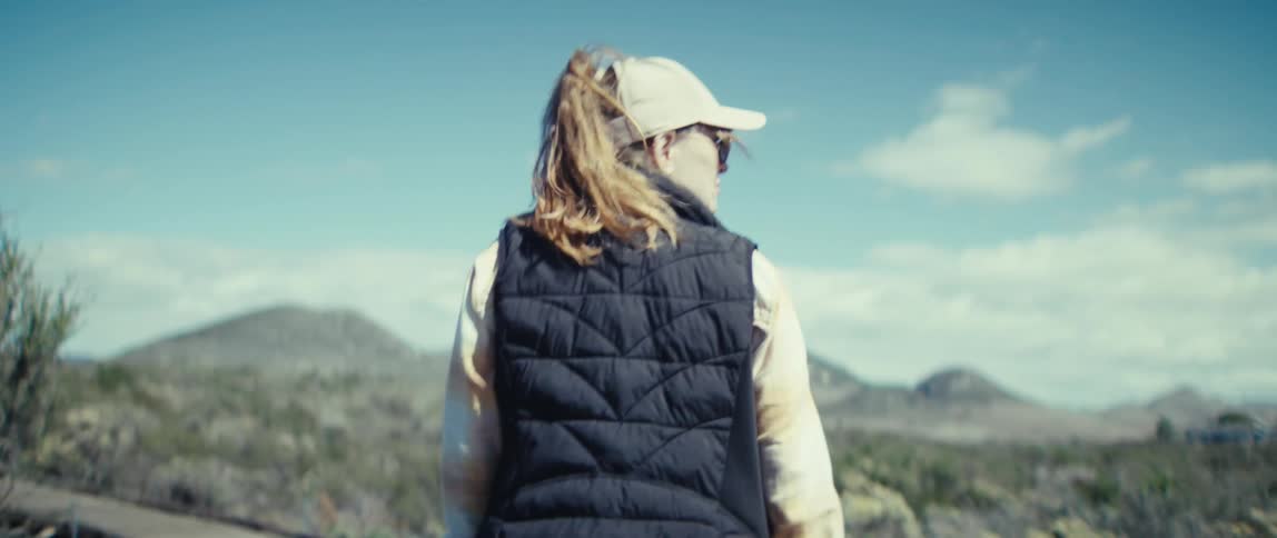 A woman walks across a grassland wearing a hat sunglasses and a jacket under the sun