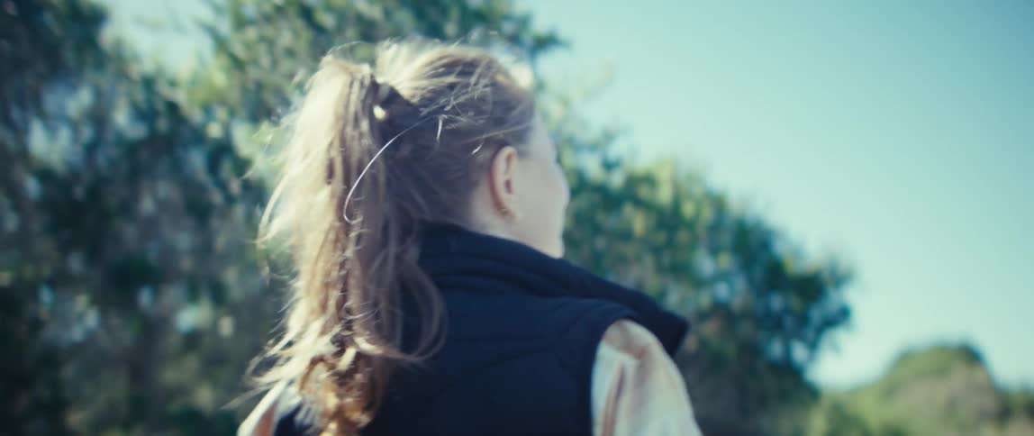 The video is a closeup of a woman in an outdoor environment with her hair tied back in a high ponytailshe is wearing a black vest over a lighter top and the sunlight is creating a bright flare from behind her