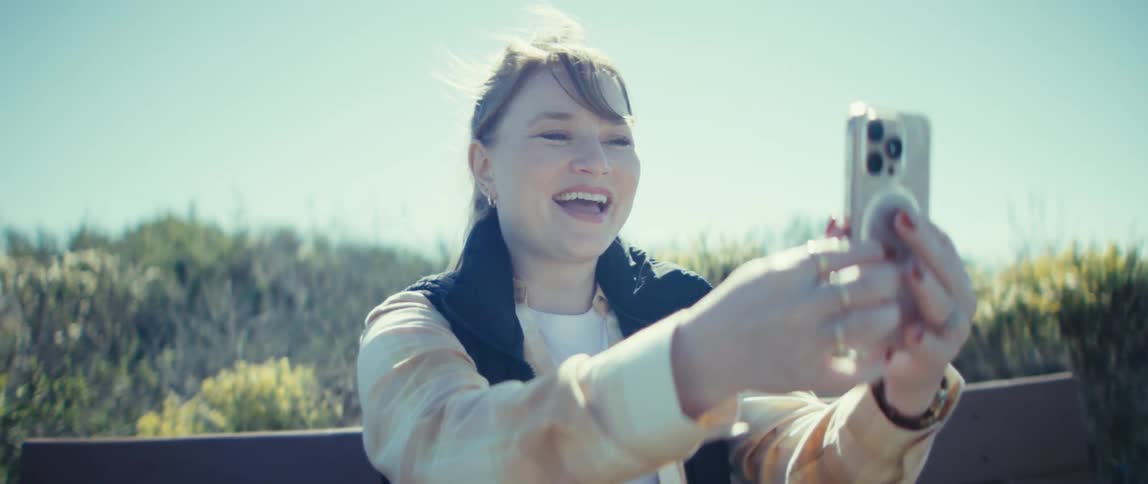 A woman sits on a wooden bench outdoors initially looking down at her phone then smiles and raises the device to begin a video call she appears happy and relaxed as she waves and engages with the person on the other end of the phone with a sunny dry