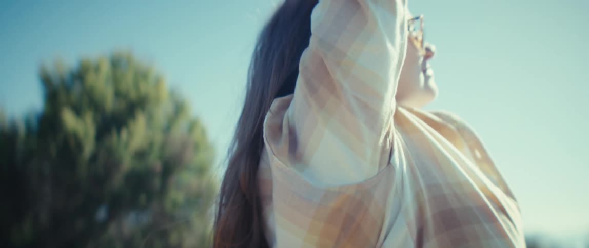 A young woman with long straight brown hair facing away from the camera raises her arms above her head while standing outside on a sunny day with a blurred mountain landscape in the background