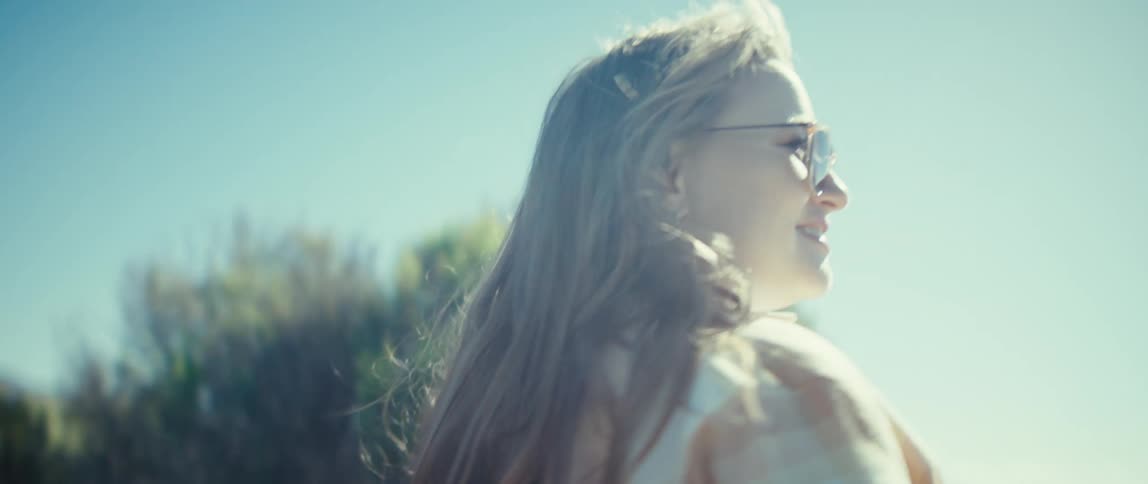 A woman wearing sunglasses smiles as she turns her head from the side to the back her hair blows in the wind and she stands outdoors against a bright blue sky and a coastal landscape