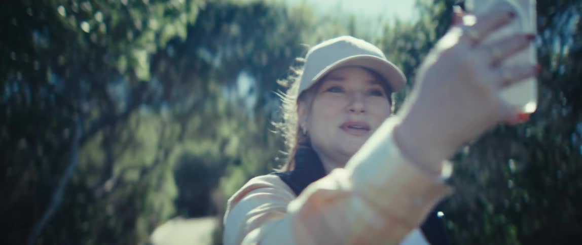 A young woman wearing a baseball hat takes a selfie video in the woods at midday