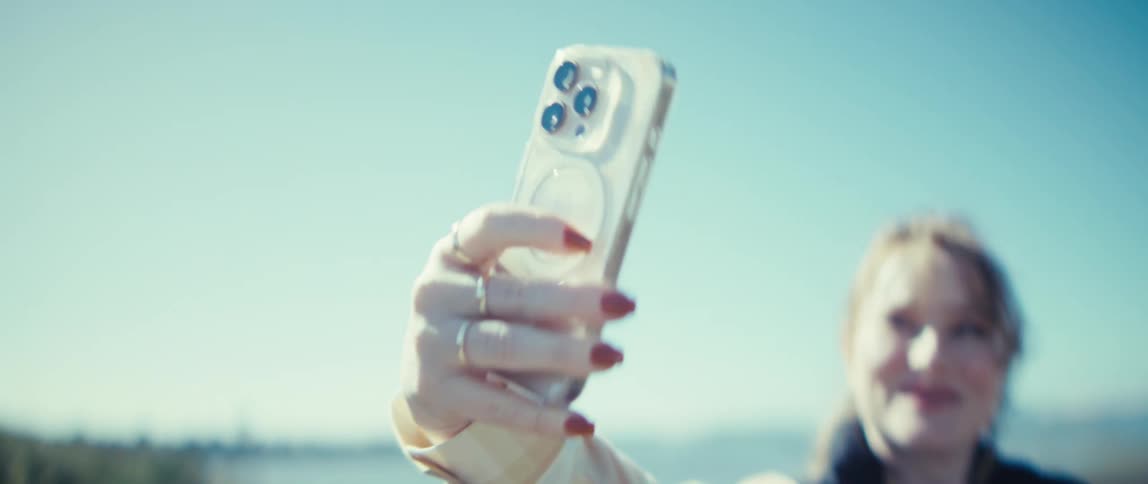 The video is a closeup of a womans hand adorned with rings and red nail polish holding a smartphone up to take a selfie or record a video outdoorsshe is smiling broadly in the background out of focus while the camera focuses on her hand and