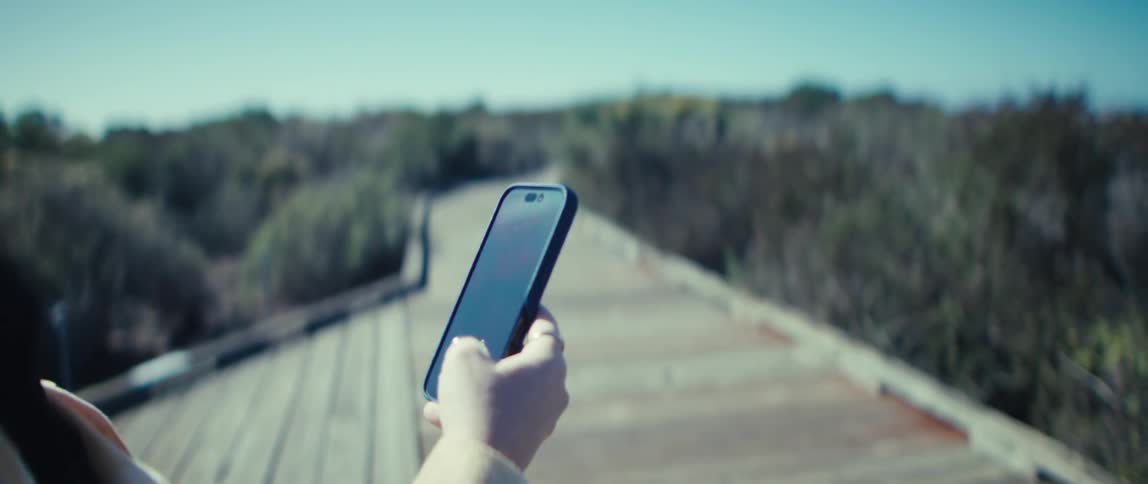 Woman holds her phone and smiles while walking along a boardwalk surrounded by vegetation