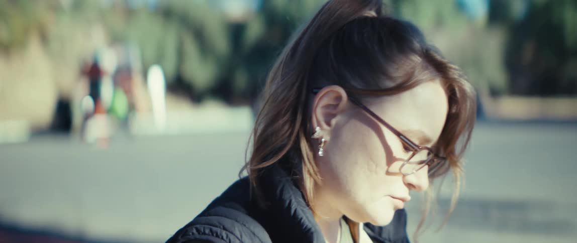 A close view shows the side profile of a woman wearing glasses a ponytail and a dark quilted vest over a tan shirt as she stands looking downward outdoors on a sunny day