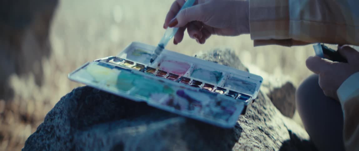 A person rubs paint from a container onto a handheld brush at midday