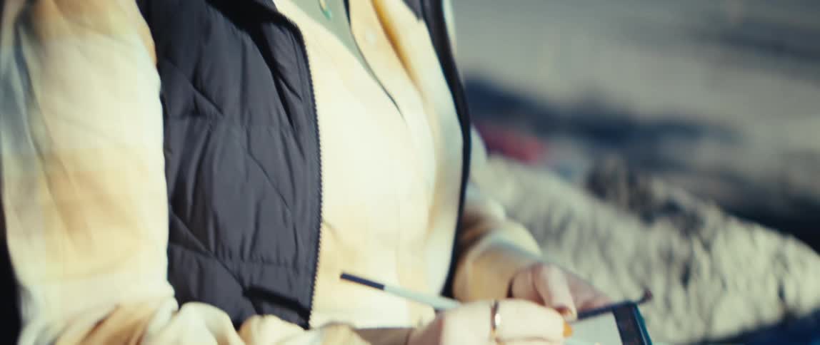 The video is a mediumclose shot of a woman with glasses and her hair partially tied back wearing a black vest over a lightcolored topit focuses on her hands using a pencil to write in a small green notebook resting on her lap suggesting she