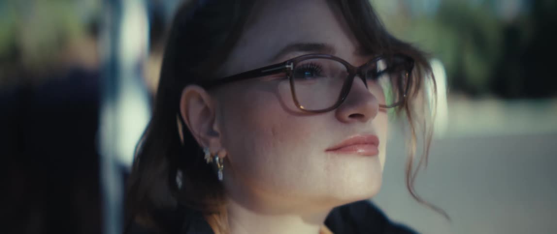 The video is a tight closeup of a womans face focused on her profile and eyesshe is wearing large darkrimmed glasses and has small hoop earrings in her ear and her expression suggests she is looking off into the distance or listening