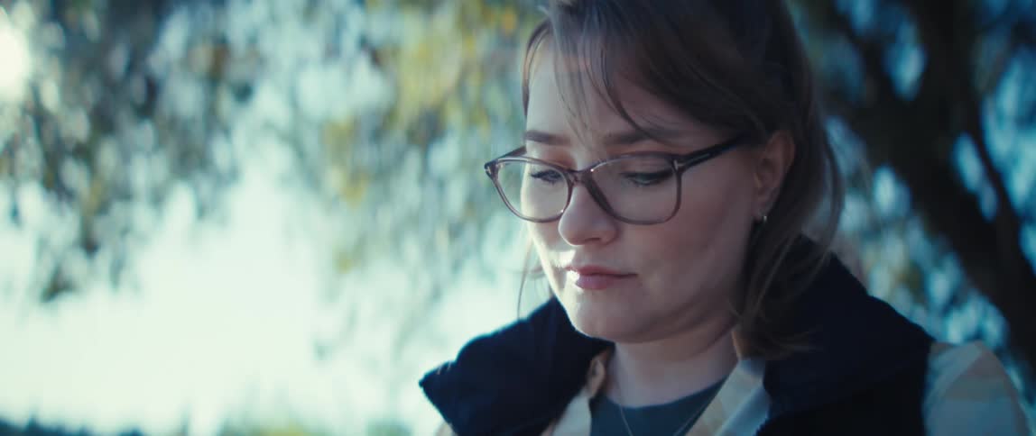 A closeup focuses on a woman outdoors wearing glasses and a jacket as she intently writes or paints in a small notebook she is holding she appears deeply focused on her task while surrounded by a leafy sunny natural environment