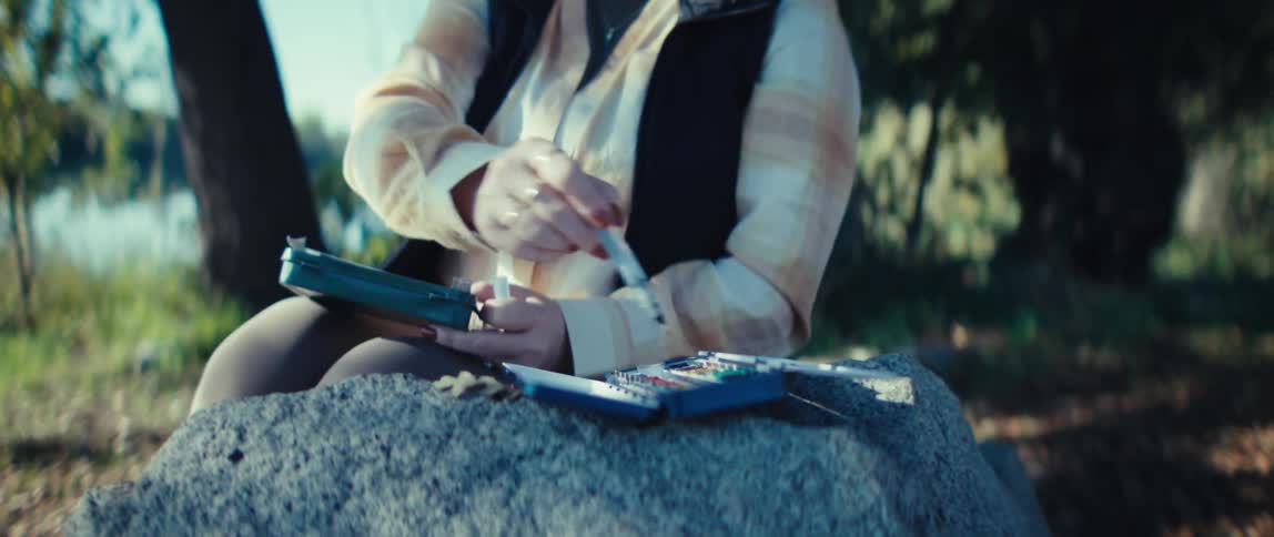 A woman sits outdoors near a body of water using a portable watercolor set to paint in a small book or on a notepad resting on her lap she is carefully mixing a color with a brush and appears focused on her artistic activity in the sunny natural enviro
