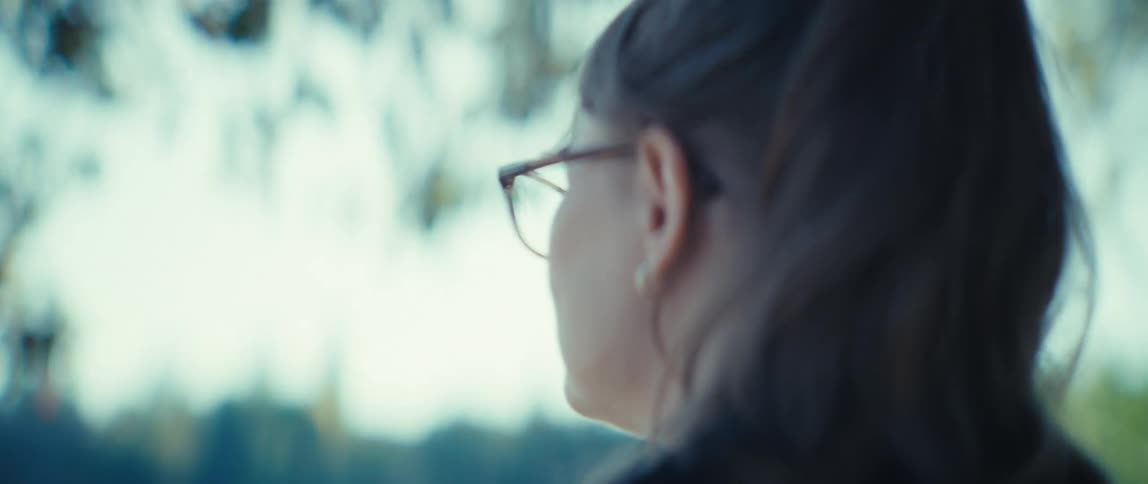 A woman with glasses and a ponytail is seen from behind outdoors with a cool blurred background