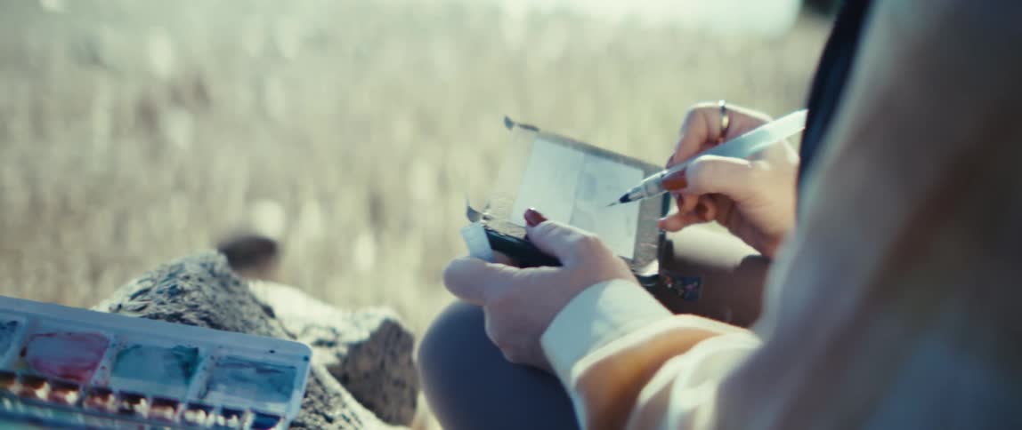 The video opens with a closeup of a persons hands mixing colors in a water paint palette resting on a rockit then shifts focus to the persons other hand as they apply paint to a small piece of paper or sketchbook they are holding suggesting