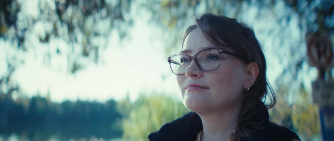 A woman wearing glasses pauses from writing in a small notebook she smiles while looking away from the camera and trees line the background next to a body of water