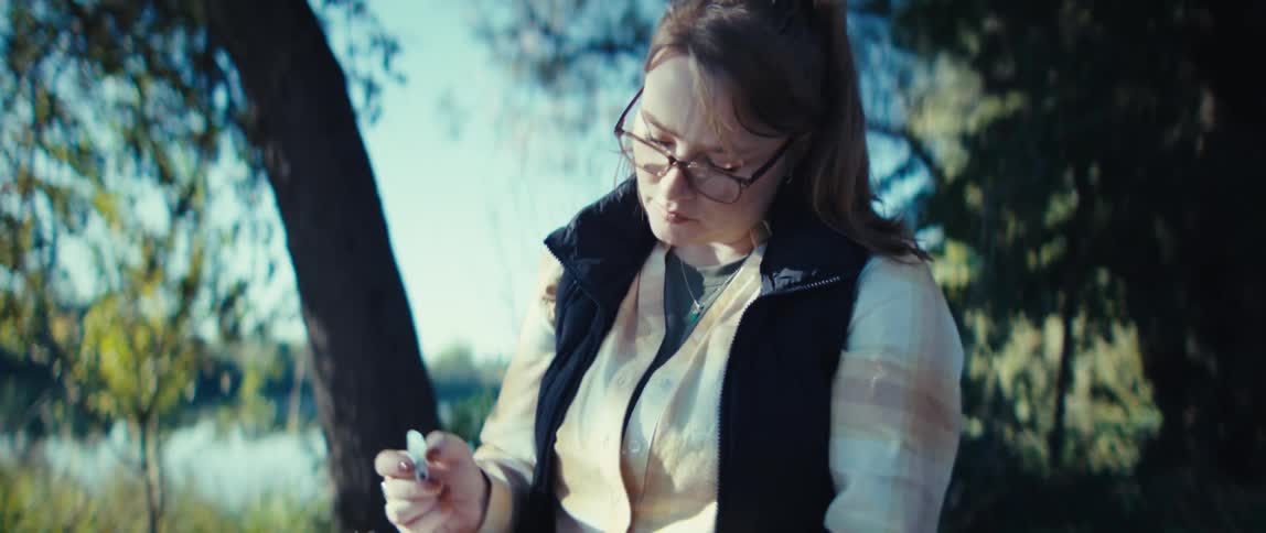 A woman sitting on a bench outdoors holding a small sketchbook and painting with a small brush