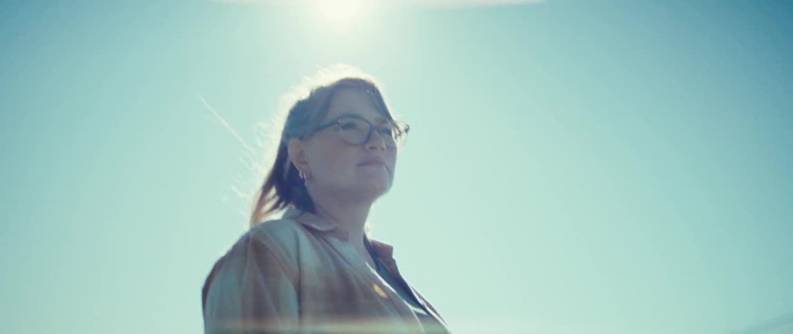 A woman wearing a plaid shirt and glasses stands outdoors looking slightly up and offcamera with a thoughtful expression the bright sun is directly behind her creating a strong backlight and lens flare against the clear blue sky