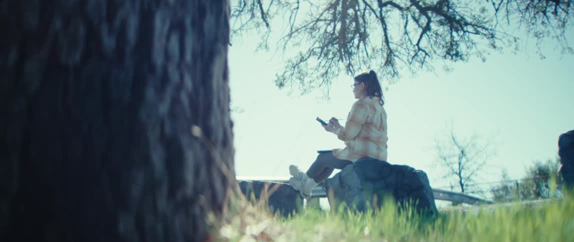 Woman sits on a rock under a trees shade while using her phone