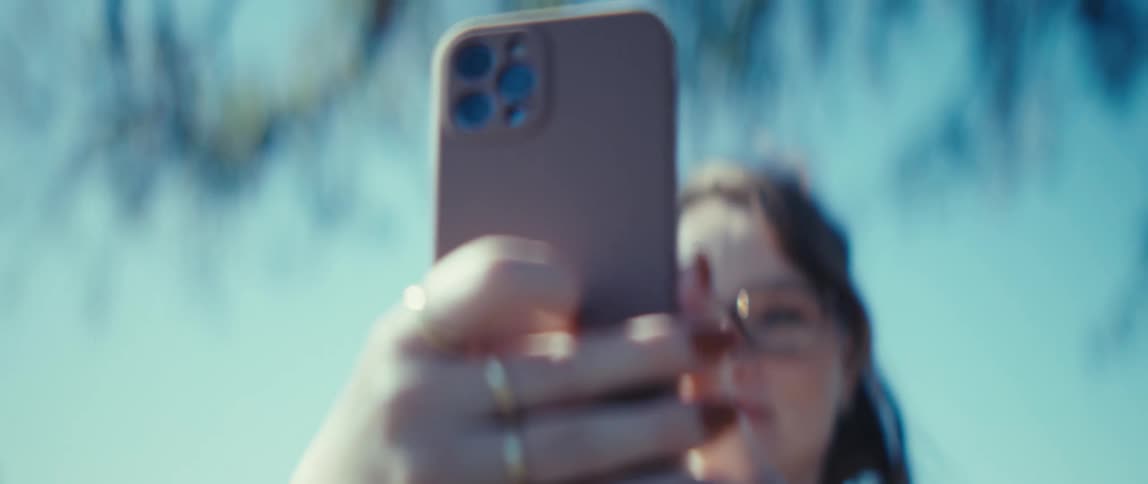 A woman wearing glasses and smiling outdoors raises her smartphone and angles it up towards the sky suggesting she is about to take a picture or record a video the shot focuses on the phones camera array and the blue sky background as the woman remains