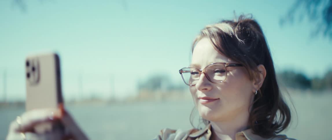 Woman with glasses smiles while holding her phone outdoors