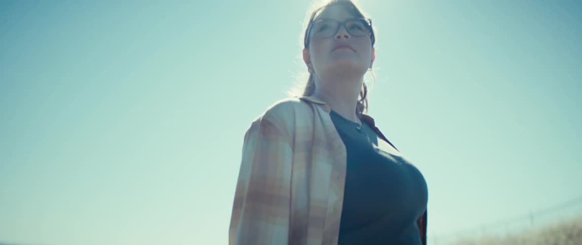 The video is a lowangle medium shot of a woman with glasses and a plaid shirt standing outdoors in a dry grassy fieldshe is looking up and away from the camera with the bright sun creating a strong lens flare over a clear sky