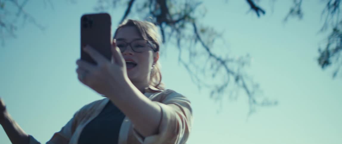 Person wearing glasses and light shirt holds phone in front of branches and bright sky