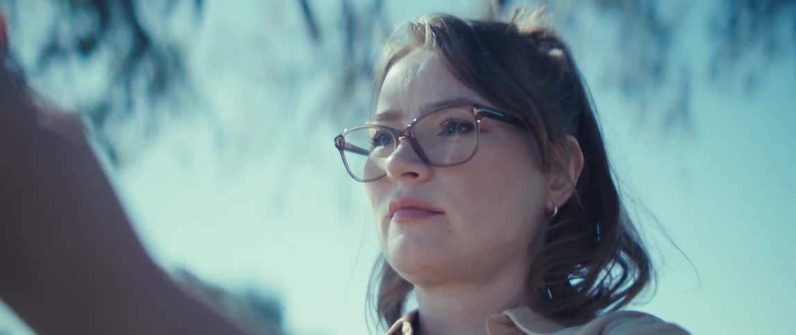 A close view shows the side profile of a young woman wearing tortoiseshell glasses with her hair partially pulled back holding up a camera or phone to take a picture on a bright sunny day outdoors