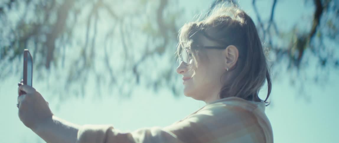 The video shows a woman outdoors facing away from the camera and holding a smartphone vertically to record or take a pictureshe turns her head back and smiles while wearing glasses continuing to hold the phone up to capture the bright sundr