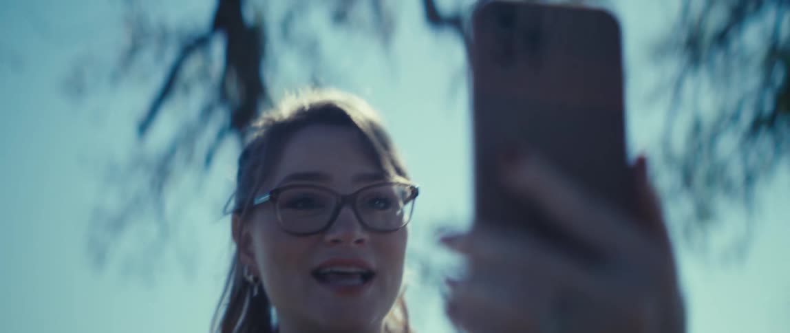 A woman wearing glasses smiles and holds a smartphone with a case in front of her while she is talking to someone by video call and the scenery is outdoors with bright sun and a tree