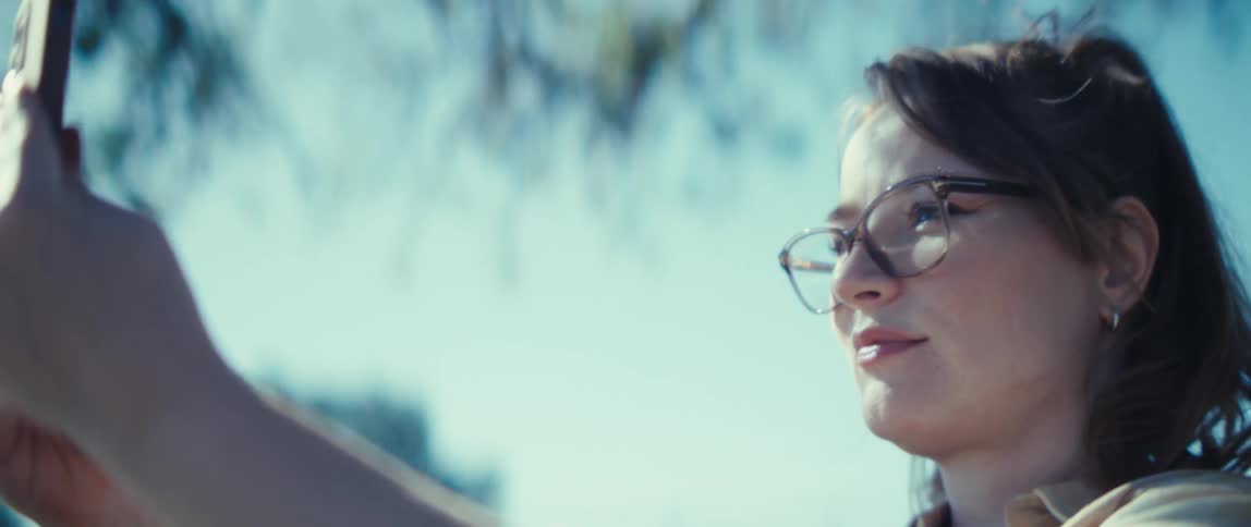 A young woman wearing glasses takes a selfie with her mobile device under a tree at midday