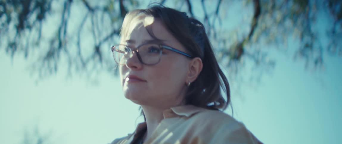 A woman wearing glasses holds a tablet in her hands she looks up and smiles at the camera and the bright sun shines through the bare branches of a tree behind her